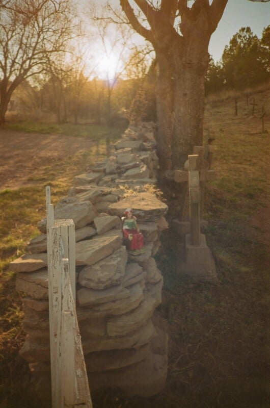 A stone wall lined with crosses runs through a grassy area at sunset. A small figurine dressed in red and green sits on the wall, and bare trees stand in the background, casting long shadows.