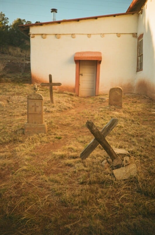 Three old gravestones, including two wooden crosses, stand on dry grass in front of a small adobe building with a white wall, a wooden door, and a window. Trees and a hill are in the background.