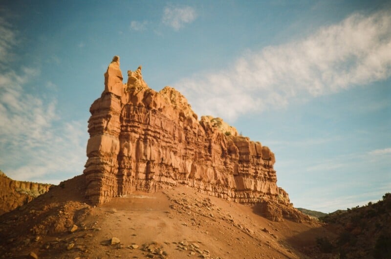 A rugged, reddish-brown rock formation stands against a blue sky with scattered clouds, surrounded by a dry, rocky landscape.