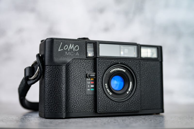 A black Lomo MC-A film camera with a blue lens sits on a gray surface against a blurred gray background. The camera features textured grips, a wrist strap, and color-coded focus distance markers.