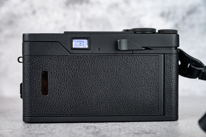 Back view of a black digital camera with a textured grip, visible viewfinder, dials, and a strap on the right side, placed on a light, speckled surface with a blurred background.