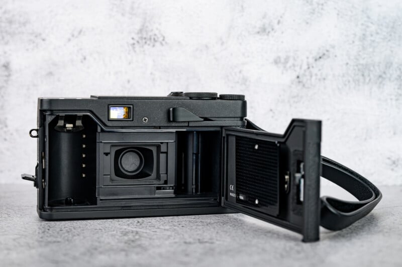 A black film camera is shown on a gray surface with its back cover open, revealing the film compartment and inner mechanisms.