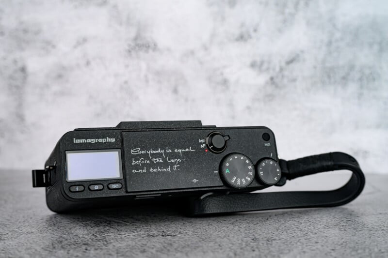 A black Lomography camera rests on a gray surface. Handwritten text on the camera reads, "Everybody is equal before the lens and behind it." The camera has a display screen, dials, and a wrist strap.