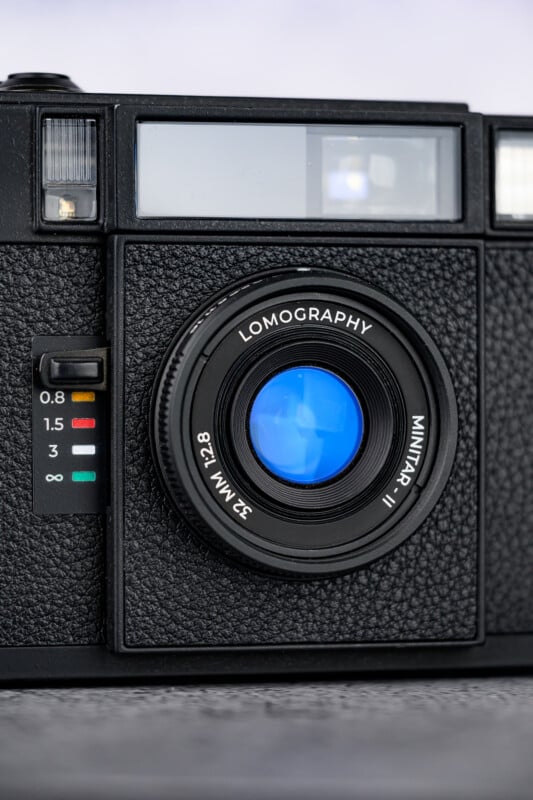 Close-up of a black Lomography camera with a textured body, showing the lens labeled “Lomography Minitar-1 Art Lens 32mm f/2.8” and aperture distance markings on the left side.