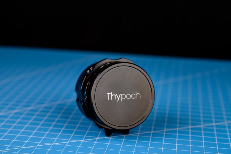 Viene con una lente de cámara "Saipoh" La tapa de la lente con la marca se asienta sobre una superficie con patrón de cuadrícula azul sobre un fondo negro.