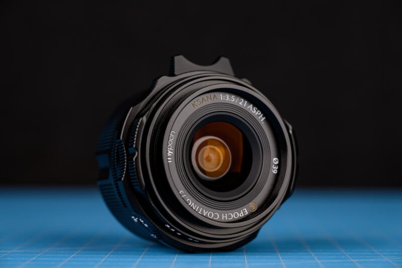 Foto de primer plano de una lente de cámara colocada sobre una superficie con patrón de cuadrícula azul, que muestra el cristal de la lente y un texto grabado que detalla el modelo y las especificaciones de la lente, sobre un fondo negro.