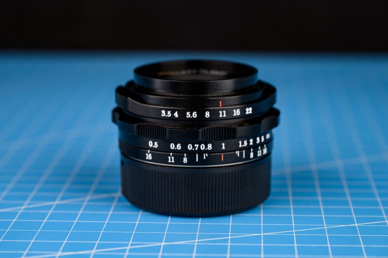 Lente de cámara negra con marcas blancas y naranjas que muestran la apertura y la distancia focal colocada verticalmente sobre una alfombra de corte azul con líneas de cuadrícula.
