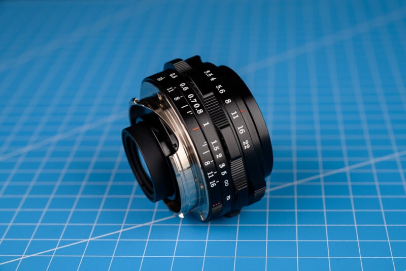 La lente de una cámara antigua con marcas de distancia y apertura se encuentra de lado sobre una superficie con patrón de cuadrícula azul.