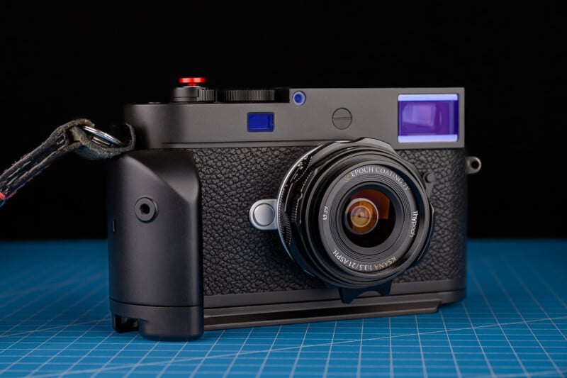 Cámara de telémetro moderna y negra con mango texturizado y lente adjunta colocada sobre una superficie con diseño de rejilla azul y fondo negro detrás.