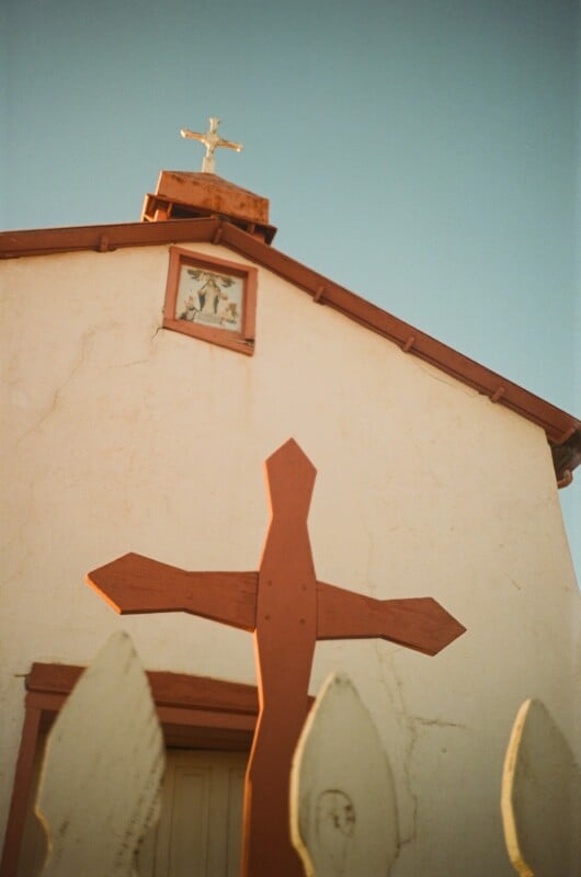 A wooden cross stands in front of a small beige church with a cross on its roof and a religious painting above the entrance, framed by a blue sky and part of a white picket fence in the foreground.