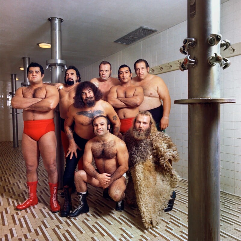 Seven muscular wrestlers in a tiled locker room, five standing and two crouching in front. Most wear red or black trunks and boots; one has long blond hair and a furry coat. Metal pipes and showers are visible in the background.