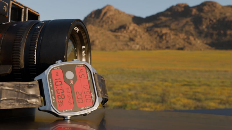 Un reloj digital con pantalla roja se coloca sobre una superficie reflectante junto a una gran lente de cámara, con las montañas y los campos al fondo borrosos bajo la clara luz del sol.