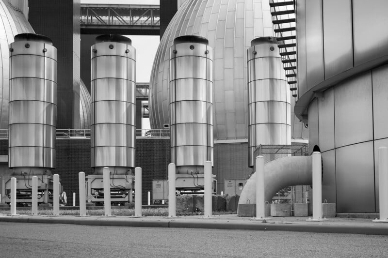 Cuatro grandes estructuras metálicas cilíndricas se encuentran frente a un edificio industrial circular en forma de cúpula, con tuberías, vallas y columnas claramente visibles; la escena es en blanco y negro.