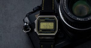 A Casio A168WE digital wristwatch with a black strap rests on top of a vintage film camera with a 50mm lens, both on a dark grey surface. The watch display shows the time as 7:07:43 and the date as Saturday the 13th.