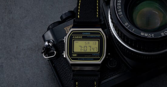 A Casio A168WE digital wristwatch with a black strap rests on top of a vintage film camera with a 50mm lens, both on a dark grey surface. The watch display shows the time as 7:07:43 and the date as Saturday the 13th.
