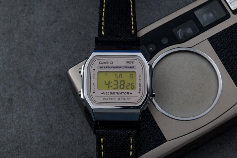 Reloj digital Casio con correa negra colocado en una cámara antigua. La pantalla del reloj muestra la hora 4:38:26 y la fecha del sábado 8. El reloj tiene etiquetas 