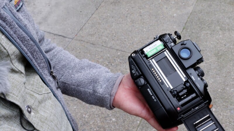 A person wearing a gray jacket holds an open film camera outdoors, showing the camera’s back with the film chamber and settings display visible. Concrete pavement is seen in the background.