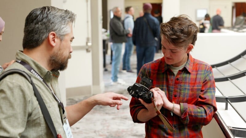 Two people stand indoors; one points at a camera while the other holds and inspects it. Both seem focused on the camera. Several people are blurred in the background in a hallway.