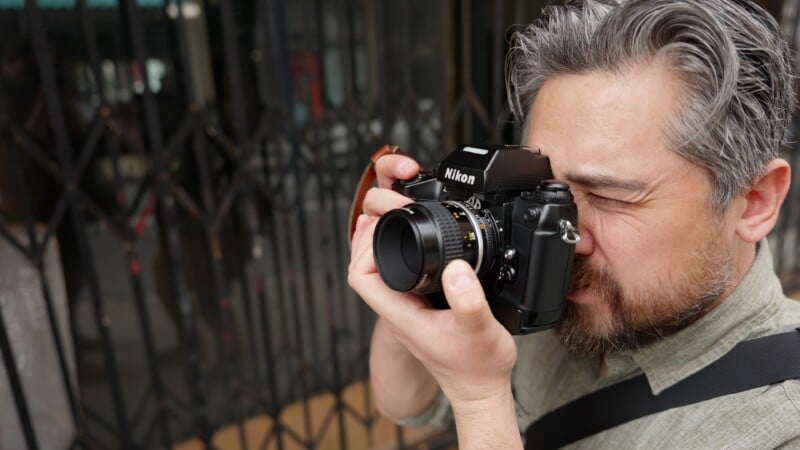 A man with gray hair and a beard is holding a Nikon camera up to his eye, appearing to take a photo. He is standing outside in front of a metal gate and wearing a light green shirt.