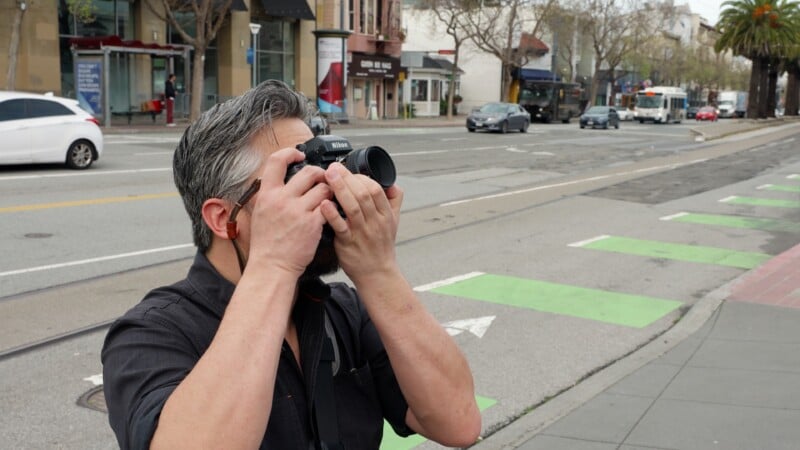 A man with gray hair and a beard stands on a city sidewalk, holding a Nikon camera up to his face as he takes a photo. Cars, shops, and trees are visible in the background along the street.