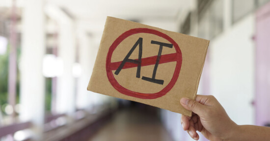 A hand holds a cardboard sign with the letters "AI" inside a red circle with a diagonal line through it, indicating opposition to artificial intelligence, in a blurred indoor setting.