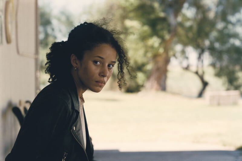 Una mujer joven con el pelo rizado y una chaqueta negra se sienta al aire libre y mira pensativamente a la cámara. El fondo está borroso y muestra árboles verdes y luz solar.