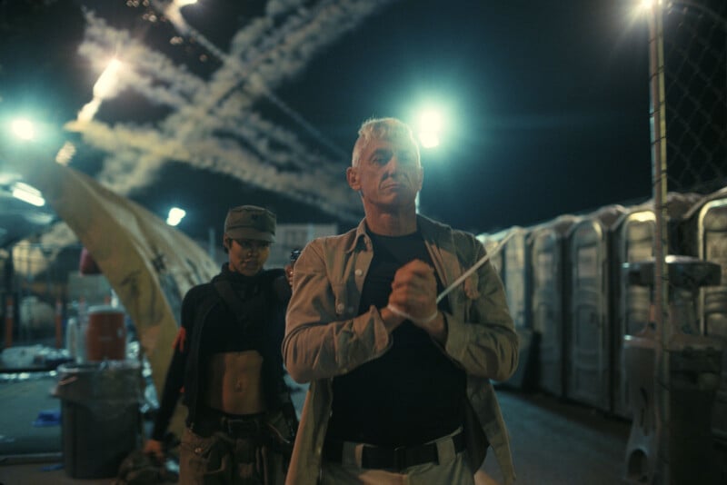 Un hombre de cabello gris, con las manos atadas con cremalleras, camina resueltamente en la noche, seguido por una mujer con uniforme militar y expresión seria; luces brillantes y rayas iluminan el cielo.