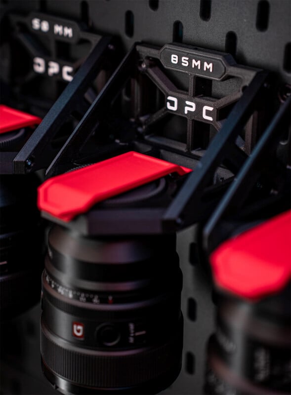 Primer plano de la lente de la cámara montada en un soporte de pared con la etiqueta "85MMOPC," Se ven componentes negros y rojos, otras tomas aparecen borrosas en el fondo.