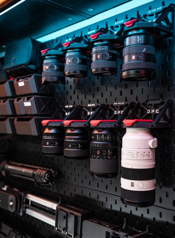 El tablero perforado montado en la pared muestra múltiples lentes de cámara, estuches para lentes y accesorios de cámara cuidadosamente dispuestos en estantes y ganchos. La configuración tiene un aspecto moderno y profesional y buena iluminación.