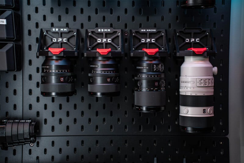 Cuatro lentes de cámara de diferentes tamaños están montados en una pared negra perforada con soportes etiquetados, incluido un teleobjetivo blanco a la derecha y tres lentes negros etiquetados 50MM, 85MM y 100 MACRO de izquierda a derecha.