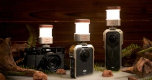 Three cameras with small, glowing lanterns mounted on top are displayed on a wooden surface, surrounded by ferns and pieces of wood, against a wooden backdrop.
