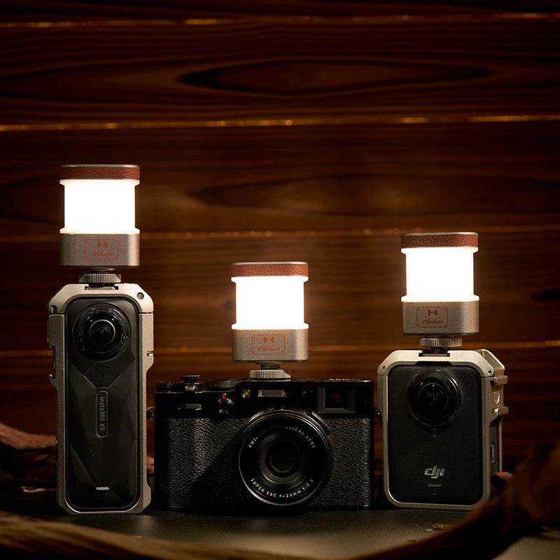 Se muestran tres cámaras sobre una superficie de madera, cada una con una pequeña luz LED cilíndrica montada en la parte superior, que proyecta una luz brillante en un ambiente cálido y oscuro con paneles de madera al fondo.