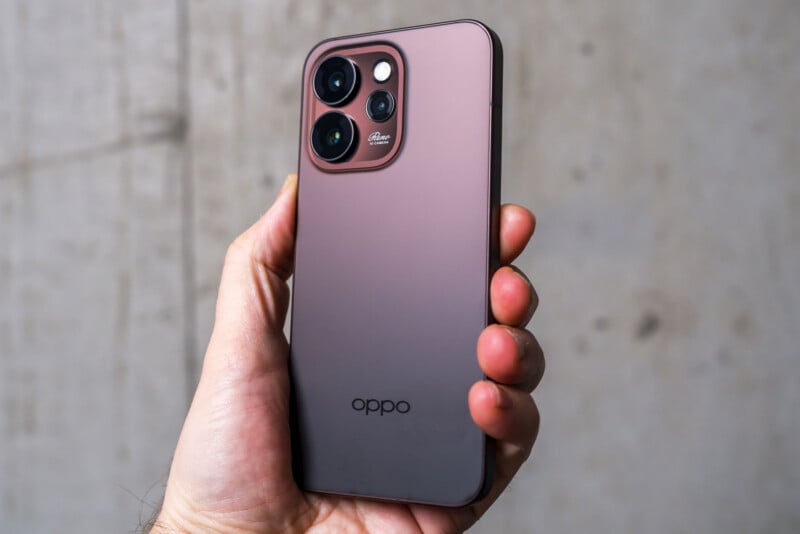 A hand holding a matte pink Oppo smartphone with a large square camera module featuring four lenses and the Oppo logo at the bottom. The background is a blurred beige wall.