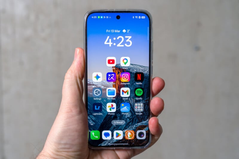 A hand holding a smartphone displaying the home screen with various app icons, including YouTube, Gmail, Netflix, Spotify, and Google apps, with the time shown as 4:23 and the date as Friday, March 13.