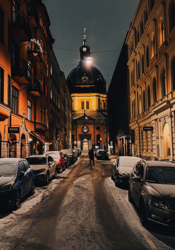 A narrow city street at night, lined with parked cars dusted with snow, leads to an illuminated domed building; a person walks down the center of the road between tall, historic buildings.
