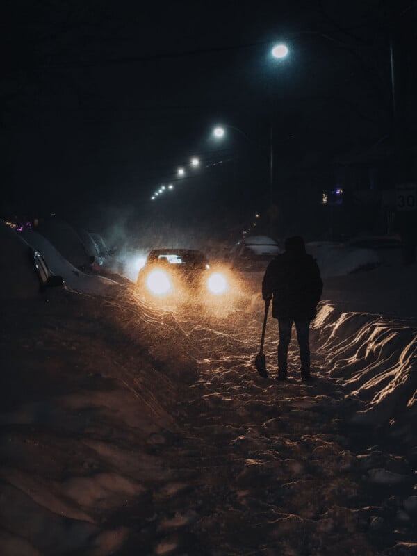 A person holding a shovel stands on a snow-covered street at night, illuminated by the headlights of an approaching car and streetlights, as snow falls heavily.