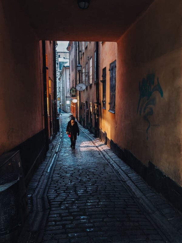 A person walks down a narrow cobblestone alleyway bordered by tall, orange buildings; warm lights illuminate the passage, and graffiti is visible on the right wall.