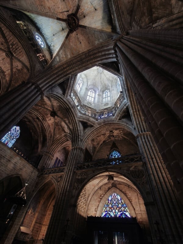 View of a grand Gothic cathedral interior, showcasing soaring stone columns, arched ceilings, and intricate stained glass windows with colorful patterns. Light filters in through the windows, illuminating the architectural details.