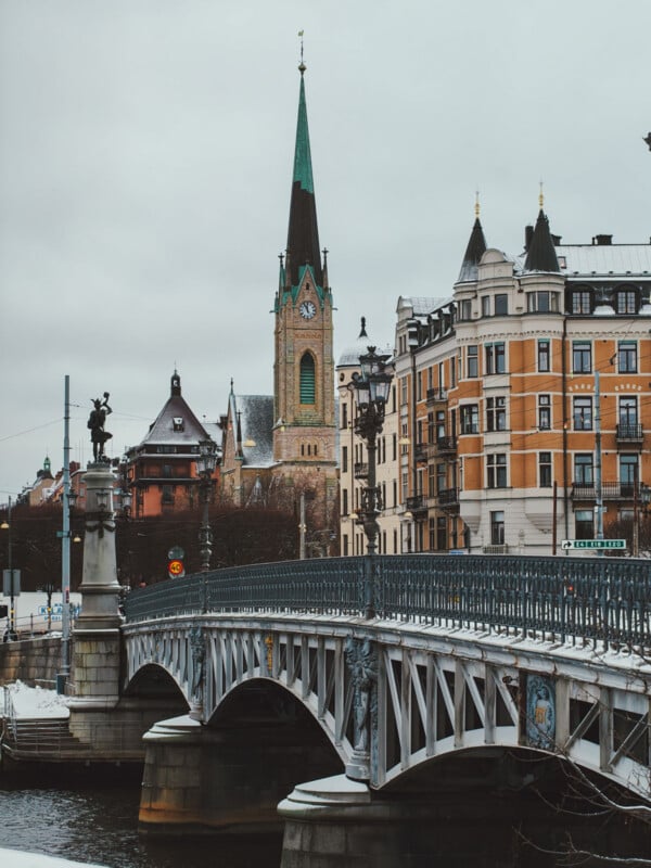 A metal bridge crosses over water in a city with ornate, historic buildings and a tall church spire in the background under a cloudy sky.