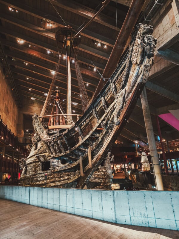 A large, well-preserved wooden ship is displayed inside a spacious modern museum with high ceilings and dramatic lighting. The ship's detailed structure and rigging are visible.