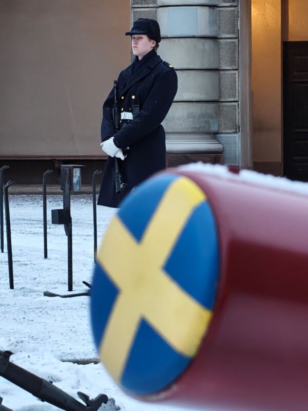 A uniformed guard stands at attention on a snowy street, with a large red object bearing the Swedish flag in the foreground and a historic stone building behind.