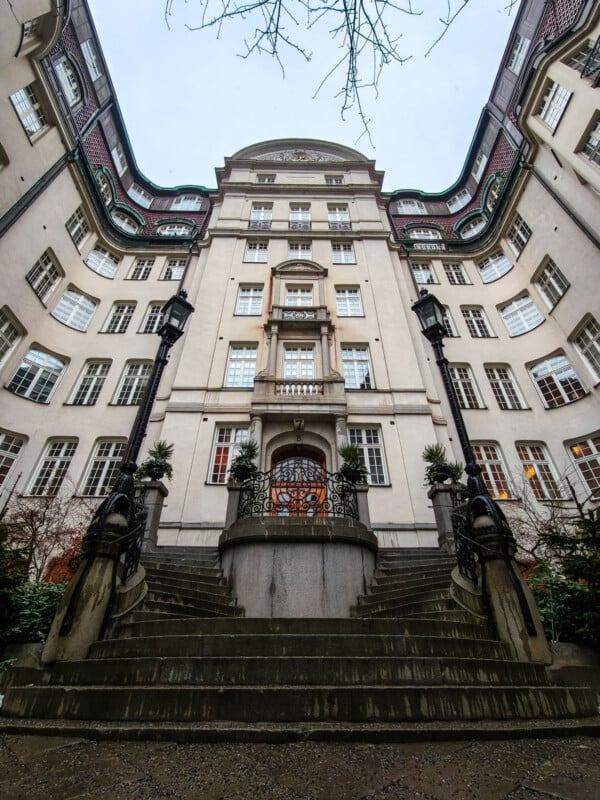 Ornate, curved stone staircase leads to the entrance of a grand, historic building with many large windows, decorative ironwork, and intricate architectural details. Leafless branches are visible above, framing the scene.