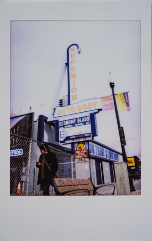Un hombre se encuentra cerca de una valla metálica frente a un edificio con grandes antigüedades "Cuerpo de alta calidad" logotipo y varios otros logotipos comerciales. Bancos y farolas visibles; cielo nublado.