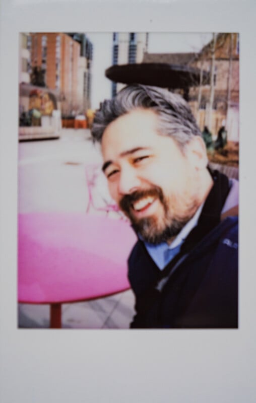 Un hombre barbudo y de pelo gris se sienta en una mesa rosa al aire libre y sonríe a la cámara. Los edificios y los árboles de la ciudad son claramente visibles en el fondo borroso.