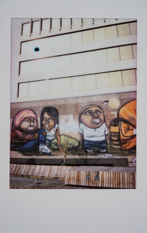 Coloridos murales en las paredes del edificio representan personajes de dibujos animados sentados o de pie, con un banco de madera y una pequeña planta frente al mural. Hay grandes ventanales encima del edificio.