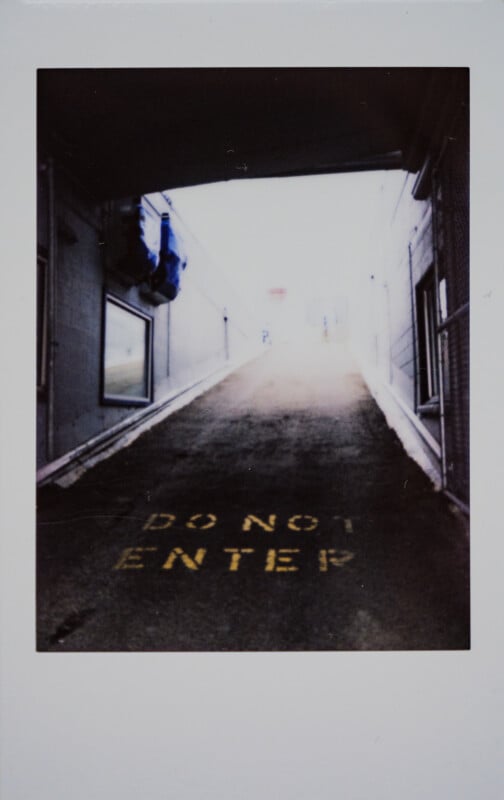 La rampa conduce hacia arriba hacia una luz brillante, "No entrar" Hay palabras amarillas pintadas en el suelo. El área está delimitada por paredes y techo, con ventanas y tuberías visibles a la izquierda.