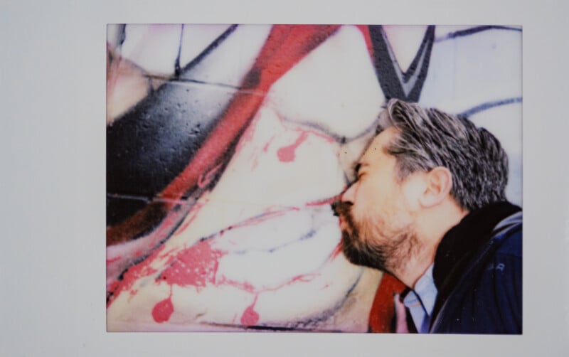 Un hombre barbudo y de cabello gris presiona su rostro contra una pared cubierta de graffiti, aparentemente interactuando con la colorida obra de arte abstracta.