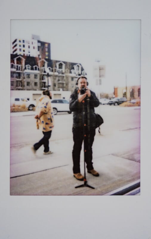 Un hombre sostiene una cámara sobre un trípode y fotografía su reflejo en una ventana de una calle de la ciudad. Otro hombre pasa al fondo. Al otro lado de la calle se ven claramente los edificios y los coches aparcados.