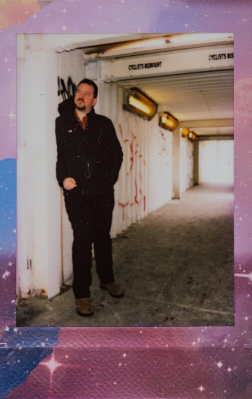 Un hombre vestido de oscuro se apoya contra una pared cubierta de graffiti en un túnel bien iluminado que dice "metro ciclista," El borde presenta un diseño espacial colorido, parecido a un cielo estrellado.