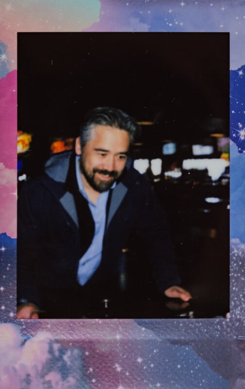 Un hombre barbudo y de pelo gris, vestido con una chaqueta oscura y una camisa azul, se inclina hacia adelante y sonríe. Esta foto tiene un borde colorido con nubes y estrellas, lo que le da un aspecto de ensueño con temática espacial.